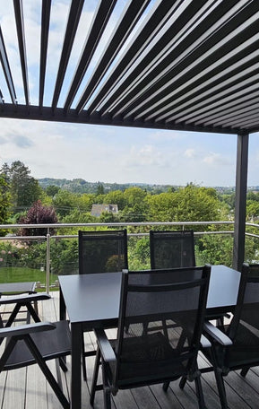 Anthrazitfarbene Pergolux Pergola auf einer Terrasse mit Glasgeländer und Esstisch, mit Blick auf grüne Landschaft.