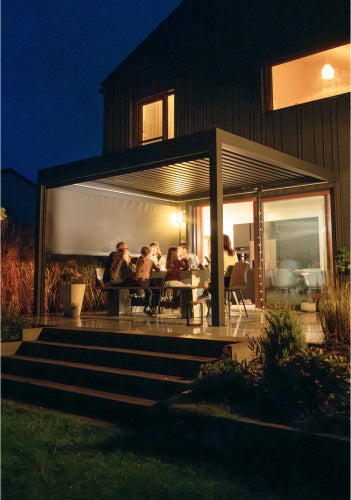 Beleuchtete Pergolux Pergola am Abend mit Personen, die gemeinsam unter dem Lamellendach sitzen und den Abend genießen.