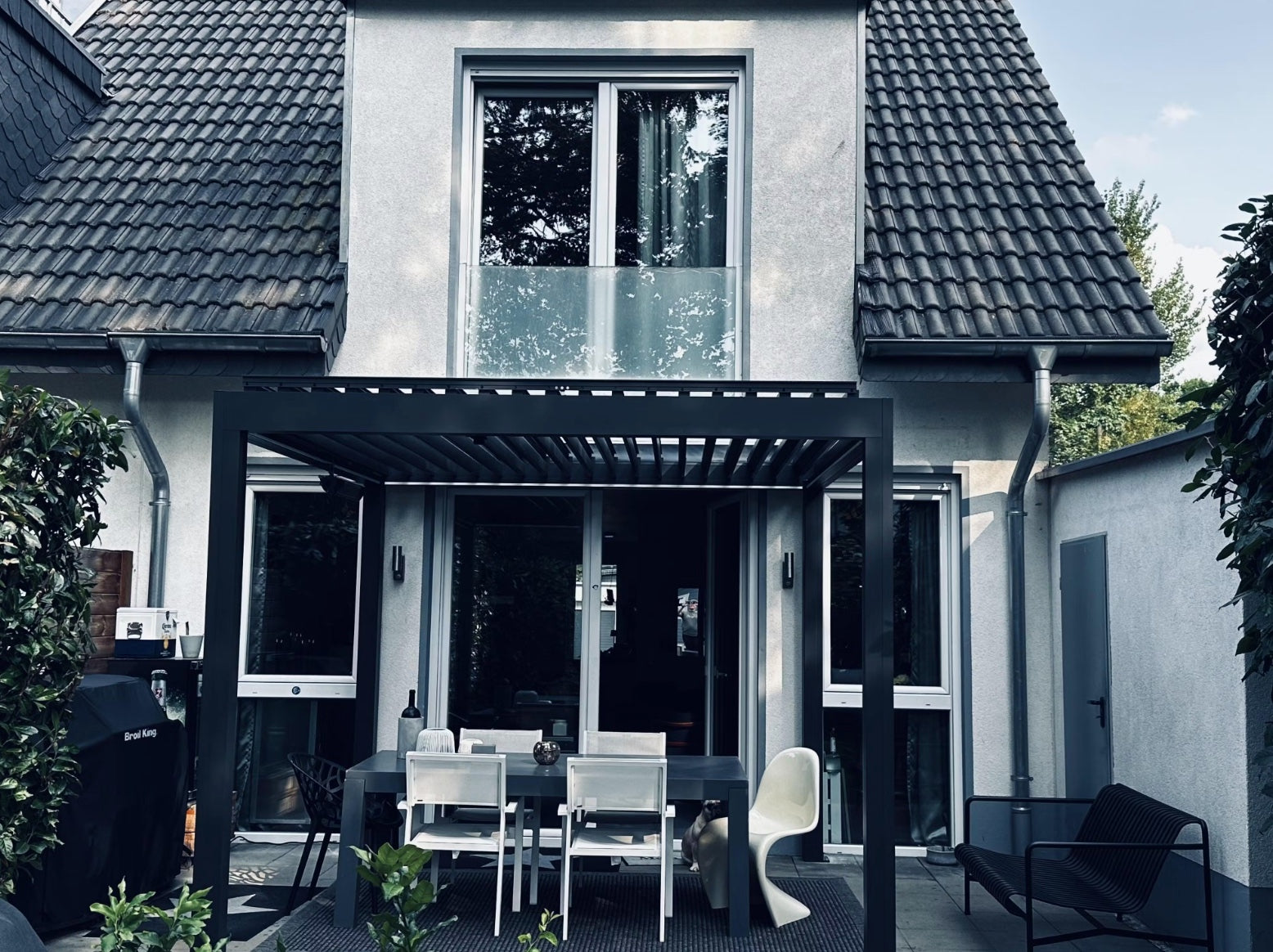 Anthrazitfarbene Pergolux Pergola als Terrassenüberdachung an einem Einfamilienhaus mit Essbereich und Grill.