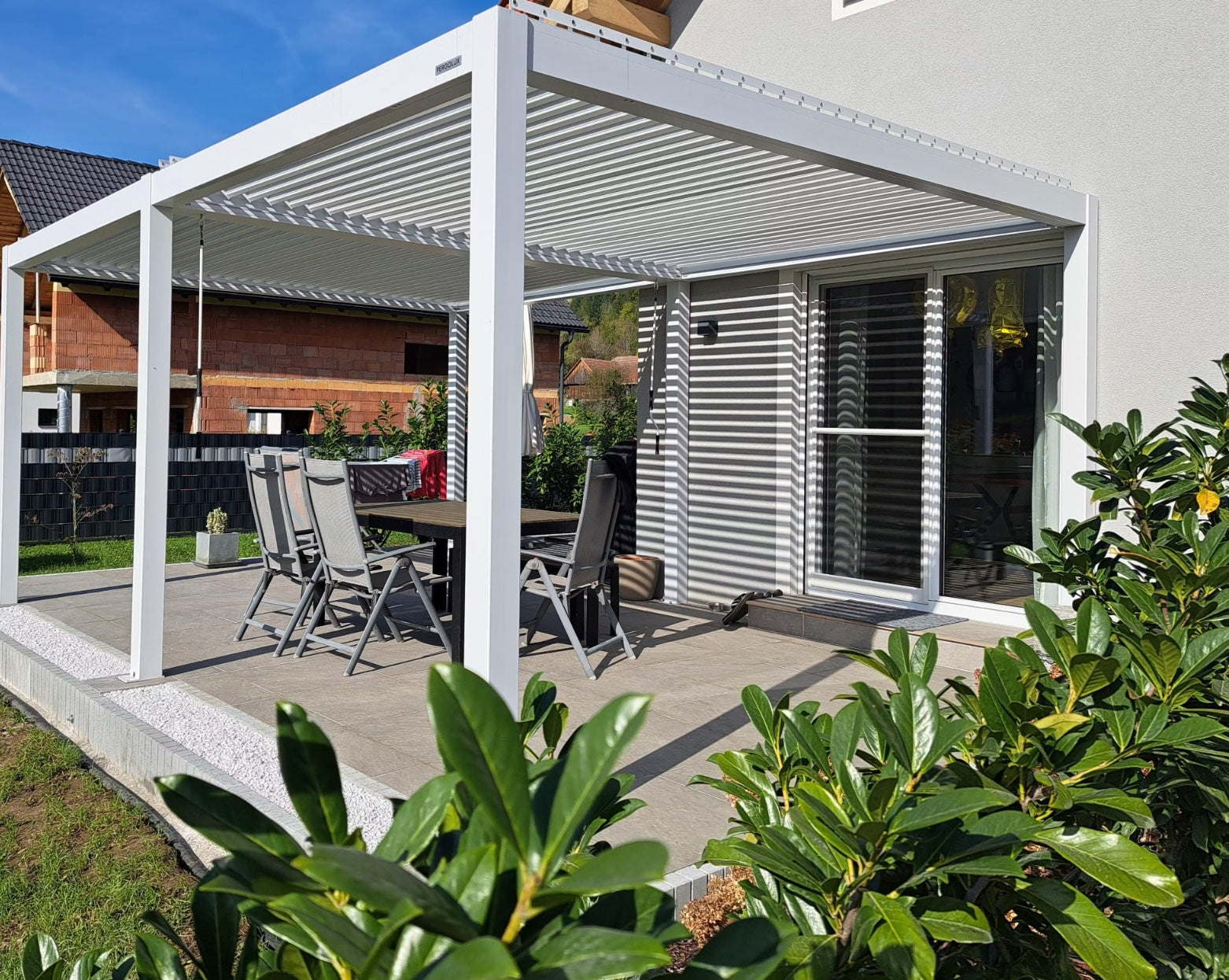 Weiße Pergolux Pergola mit heruntergelassenem Screen-Rollo und beleuchteter Lounge mit Feuerstelle bei Dämmerung.