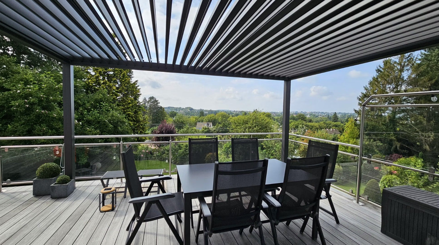 Anthrazitfarbene Pergolux Pergola auf einer Terrasse mit Glasgeländer und Esstisch, mit Blick auf grüne Landschaft.
