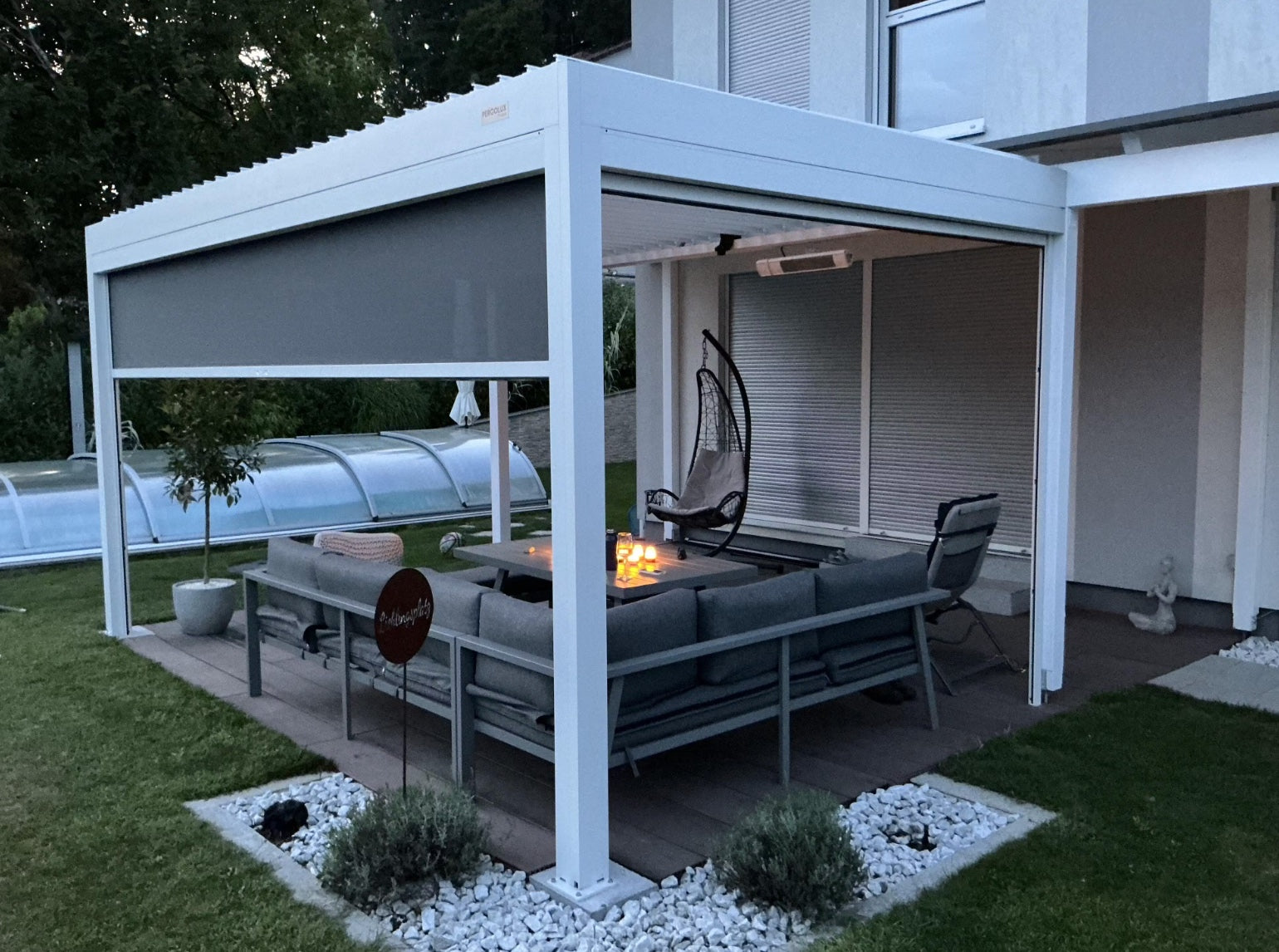 Weiße Pergolux Pergola mit heruntergelassenem Screen-Rollo und beleuchteter Lounge mit Feuerstelle bei Dämmerung.